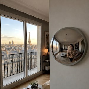 May include: A circular convex wall mirror with a smooth, light-coloured frame mounted on a wall. The mirror reflects a room interior, including a person holding a smartphone, while the adjacent window shows a view of the Eiffel Tower in Paris.