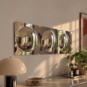 May include: Three square wall mirrors with rounded corners and circular reflective centres, featuring a polished silver finish. The mirrors are mounted horizontally in a row, reflecting the interior room space and natural light.