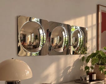 Pode incluir: Três espelhos de parede quadrados com cantos arredondados e centros refletores circulares, com acabamento em prata polida. Os espelhos estão montados horizontalmente em fila, refletindo o espaço interior e a luz natural.