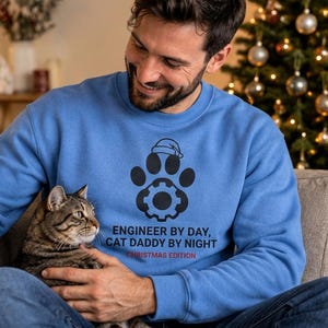 May include: A blue crewneck sweatshirt featuring a black graphic of a paw print with a gear in the center and a small Santa hat on top. Below the graphic, the text reads: ENGINEER BY DAY, CAT DADDY BY NIGHT, with CHRISTMAS EDITION in red.
