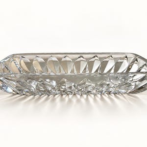 May include: A clear, rectangular glass dish featuring a faceted, geometric pattern along its exterior walls. The dish has a shallow, elongated form with beveled edges and a smooth, flat interior base, reflecting light through its transparent structure.
