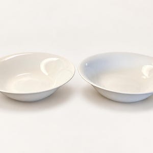 May include: Two matching white ceramic bowls with a smooth, glossy finish and a wide, shallow basin design. Each bowl features a simple, rounded rim and a clean, minimalist aesthetic, presented side by side against a plain, neutral background.