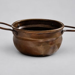 May include: A round, deep copper bowl with a dark, weathered patina and a hammered texture. The bowl features two thin, curved metal handles attached to opposite sides with small rivets, providing a functional grip for this metallic vessel.