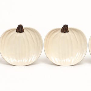 May include: A set of four off-white ceramic dishes shaped like pumpkins. Each dish features a glossy finish, vertical ridges mimicking the texture of a pumpkin, and a small, dark brown stem at the top center of the rim.