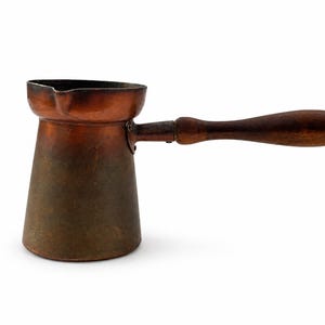 May include: A copper coffee pot with a tapered cylindrical body, a wide rim featuring a small pouring spout, and a long, dark brown wooden handle attached to the side. The metal surface shows a weathered, darkened patina throughout.