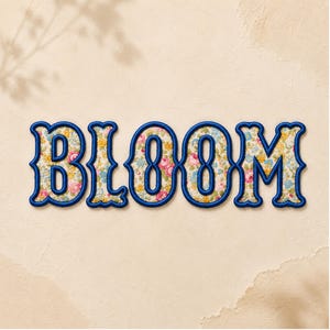 Puede incluir: La palabra BLOOM se muestra en letras grandes y decorativas con un borde grueso bordado en azul oscuro. El interior de cada letra presenta un patrón floral denso y colorido con pequeñas flores rosas, amarillas y azules sobre un fondo crema.