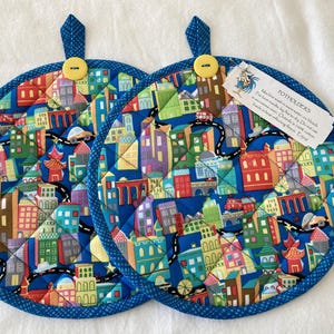 May include: Two round potholders with a vibrant city scene design. The blue fabric features colorful buildings, roads, and cars. Each potholder has a blue loop and a yellow button. The text "TOTHOLDERS" is visible on a tag.