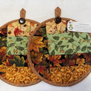 May include: Two round quilted potholders featuring a patchwork design of autumn leaves in shades of orange, yellow, green, and black. Each potholder has a brown button at the top, a hanging loop, and a tag with text including POTHOLDERS.