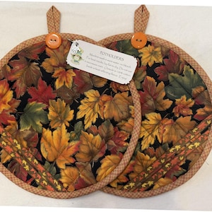 May include: Two round fabric potholders featuring a black background with a colorful autumn maple leaf print. Each has a brown checkered trim, an orange button, and a hanging loop. A white tag reads: POTHOLDERS Machine wash in warm water, no bleach. For best results, lay flat to dry. Do not use in microwave oven. Outside is 100% cotton. Inside a heat reflecting fleece. Enjoy!