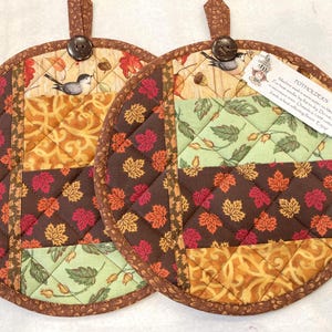 May include: Two round quilted potholders featuring a patchwork design of autumn leaves, birds, and floral patterns in shades of brown, orange, and green. Each potholder has a brown fabric loop with a dark button and a white care instruction tag.