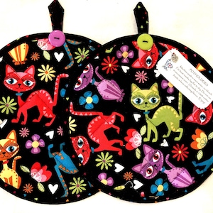 May include: Two round fabric potholders featuring a black background with a colorful pattern of stylized cats, flowers, and hearts. Each potholder has a fabric hanging loop with a button and a white tag with text including the word POTHOLDERS.