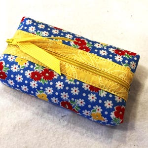 May include: A rectangular fabric pouch featuring a blue background with a repeating pattern of small white daisies and larger red and yellow flowers. A yellow zipper runs across the top, accented by a matching yellow ribbon pull tab.