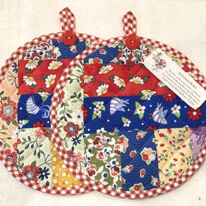 May include: Two round quilted potholders featuring a patchwork design of floral and bird patterns in red, blue, green, and yellow. Each has a red button, a red and white gingham hanging loop, and a matching gingham border. A tag includes care instructions.