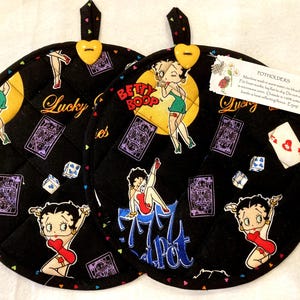 May include: Two round black quilted potholders featuring a Betty Boop print with playing cards, dice, and the text Lucky and Jackpot. Each potholder has a yellow heart-shaped button at the top loop and a white care instruction tag attached.