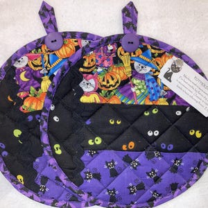 May include: Two round quilted potholders featuring a Halloween print with black cats, orange pumpkins, and purple accents. Each potholder has a purple fabric loop with a purple button and a white tag with black text detailing care instructions.