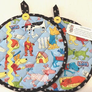 May include: Two round quilted potholders with a light blue background featuring a colorful cartoon dog print. Each potholder has a black trim with white paw prints, a yellow button, a hanging loop, and a yellow rickrack trim on one side.