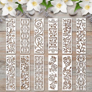May include: A set of twelve rectangular white stencils featuring various intricate laser-cut patterns, including floral motifs, geometric squares, swirling vines, and symmetrical filigree designs, arranged in two rows of six on a wooden surface.