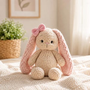 Peut inclure: Une peluche crochetée en forme de lapin avec un corps couleur crème et de longues oreilles roses texturées. Le lapin présente un petit nœud rose attaché à une oreille, des yeux brodés noirs et un détail de nez et de bouche cousus en rose.