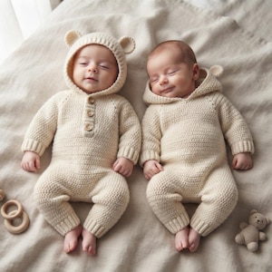 May include: Two infants wearing matching cream-colored, long-sleeved, hooded knit rompers. The left romper features a hood with small bear ears and three wooden buttons down the front chest. Both garments have ribbed cuffs at the wrists and ankles.