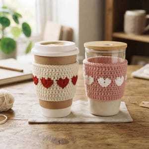 Può includere: Due tazze riutilizzabili con manicotti all'uncinetto. La tazza a sinistra è color cuoio con un manicotto color crema decorato con cuori rossi. La tazza a destra è in vetro trasparente con coperchio in bambù e manicotto rosa antico con cuori bianchi.