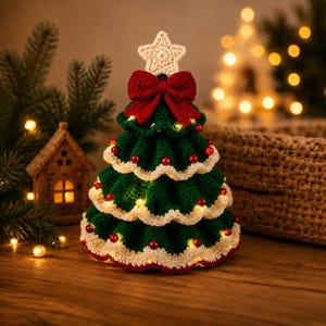 Op de afbeelding: Een gehaakte kerstboomdecoratie in donkergroen met crèmekleurige geschulpte randen, versierd met kleine rode kralen en geïntegreerde warmwitte kerstverlichting. Een grote rode gehaakte strik zit onder een crèmekleurige gehaakte stertop.