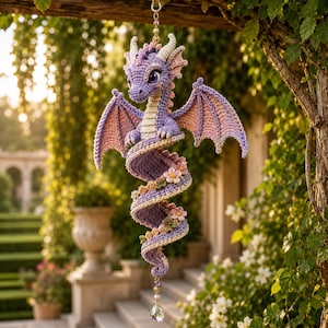 Peut inclure: Un ornement de dragon crocheté violet et rose avec une queue en spirale, orné de petites fleurs en tissu le long du corps, des cornes crème et un pendentif en cristal transparent suspendu par une chaîne dorée et un maillon perlé.