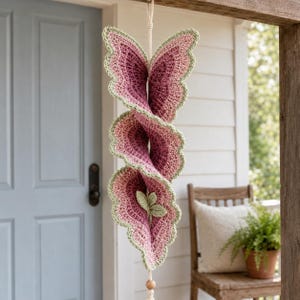 May include: A hanging crochet spiral decoration featuring layered, ruffled petal shapes in shades of dusty rose and deep magenta, finished with a light sage green scalloped edge. The structure is suspended by a cream-colored cord with a wooden bead.
