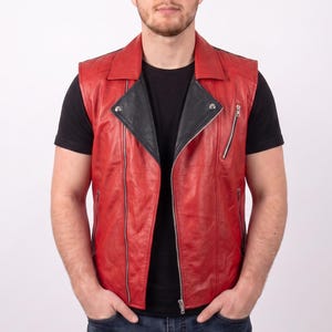May include: A sleeveless red leather vest featuring a black lapel, a silver-toned front zipper closure, and an additional diagonal zippered pocket on the upper left chest. The vest is worn over a plain black t-shirt and dark blue denim jeans.