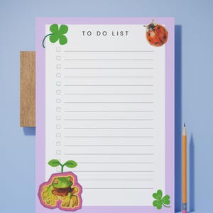 Puede incluir: Un bloc de notas rectangular para listas de tareas con un borde morado claro y centro blanco. El texto TO DO LIST está impreso en la parte superior. Las ilustraciones incluyen un trébol de cuatro hojas verde, una mariquita roja y una rana.