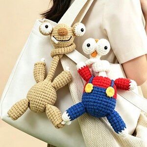 May include: Two crocheted plush figures attached to a white tote bag. One figure is tan with large eyes and a long neck. The second figure has a white head, red shirt, blue overalls with yellow buttons, and white hands, both featuring long necks.