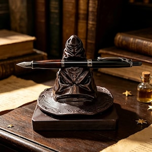 May include: A dark brown, sculpted resin pen holder shaped like a weathered, pointed hat with a wide brim, resting on a square base. A black and dark wood fountain pen with a silver clip and nib is positioned horizontally across the hat's front.
