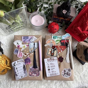 Könnte beinhalten: Zwei rechteckige, in braunes Papier eingewickelte Pakete, die mit verschiedenen Aufklebern, einem Lesezeichen mit Quaste und einem Stift verziert sind. Ein Paket enthält einen lila Stift und eine schwarze Quaste, das andere einen weißen Stift und eine weiße Quaste.