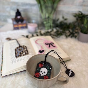 Può includere: Un segnalibro rettangolare nero con un'illustrazione di un teschio bianco circondato da rose rosse e foglie verdi, attaccato a un cordino nero con un piccolo ciondolo a forma di spada nera. Il segnalibro è inserito in una tazza di ceramica beige.