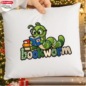 Peut inclure: Un coussin carré blanc orné d'une broderie représentant un ver de terre vert portant des lunettes noires et lisant un livre bleu. Une pile de livres rouge, orange et vert se trouve à côté du ver. Le mot "bookworm" est écrit en lettres colorées.