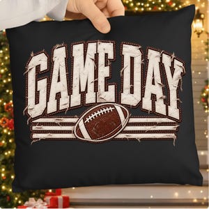 Peut inclure: Un coussin carré noir avec le texte blanc GAME DAY dans une police de style cousu et vieilli. Sous le texte se trouve un ballon de football américain marron centré entre trois lignes horizontales blanches à l'aspect effiloché et usé.