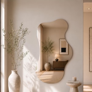 May include: A large, frameless wall mirror with an irregular, organic, wavy shape. The reflective surface displays a room interior, including a potted plant, a wooden console table, and a lamp. The mirror is mounted on a plain, light-colored wall.