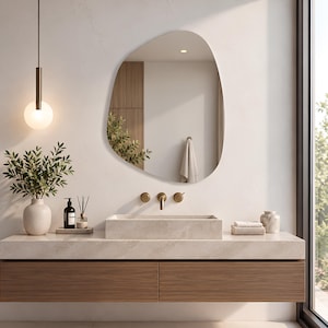 May include: An asymmetrical, organic-shaped wall mirror with a smooth, frameless edge. The mirror reflects a bathroom interior, including a light-colored stone countertop, a rectangular stone sink, brass fixtures, and a hanging pendant light.