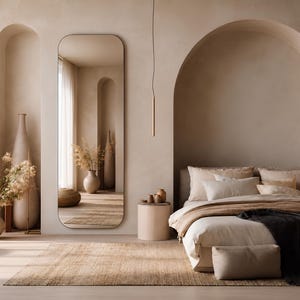 May include: A tall, rectangular floor mirror with rounded corners, featuring a thin frame. The mirror reflects a bedroom interior with arched wall niches, beige walls, a bed with neutral bedding, a dark throw, and decorative vases with dried plants.