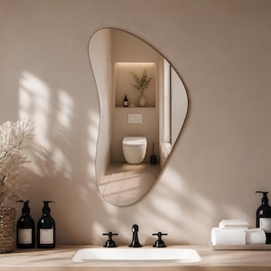 Op de afbeelding: Een asymmetrische wandspiegel met een organische vorm en een dun frame, gemonteerd boven een badkamermeubel. De spiegel reflecteert een modern badkamerinterieur met een wit toilet, een nis met een plant en een muur in neutrale tinten.