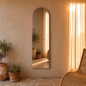 Peut inclure: Un grand miroir mural toute hauteur avec un sommet en arc arrondi. Le miroir présente un cadre fin de couleur neutre et reflète un espace intérieur ensoleillé contenant des plantes en pot et des rideaux transparents, offrant un reflet clair.