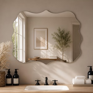 Op de afbeelding: Een wandspiegel met een golvende, geschulpte rand. De spiegel reflecteert een kamer met een houten bankje, een ingelijste print en een kamerplant. De spiegel hangt boven een badkamermeubel met een witte wastafel en een zwarte kraan.
