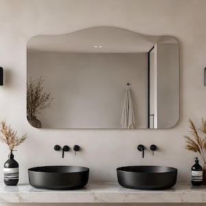 May include: A wall-mounted mirror with a soft, wavy top edge and rounded corners. Below the mirror are two matte black circular vessel sinks set into a white marble countertop, accompanied by two black wall-mounted faucets and two pendant lights.