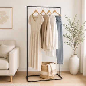 Pode incluir: Um cabideiro de metal preto com um vestido bege com decote em V, um suéter de malha bege, uma camisa branca e calças jeans azuis em cabides de madeira. Uma cesta trançada com uma manta branca com franjas repousa na base do cabideiro.