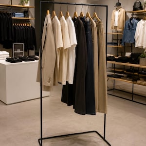 Könnte beinhalten: Ein schwarzer Kleiderständer aus Metall präsentiert eine Auswahl an Kleidungsstücken auf Holzbügeln, darunter ein beiger Blazer, weiße T-Shirts, ein dunkelbrauner Mantel, eine dunkelblaue Jeans und eine beigefarbene Hose in einem Laden.