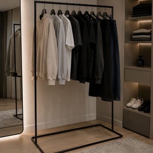 May include: A black metal clothes rail holding a row of garments on hangers, including a beige hoodie, a light grey sweatshirt, a white t-shirt, and several black shirts and trousers, all organised neatly in a sequence on the horizontal bar.