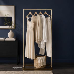 Puede incluir: Un perchero de metal dorado con un vestido beige de tirantes, un suéter de punto beige, una camisa blanca de manga larga y pantalones blancos en perchas de madera. Una cesta tejida con una manta blanca de flecos descansa en la base inferior.