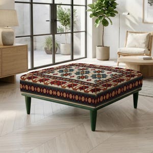 Puede incluir: Un otomán cuadrado con una parte superior de color crema con un patrón floral geométrico central en azul y rojo, bordeado por un motivo de inspiración tribal en azul oscuro y rojo. La pieza se apoya sobre cuatro patas cónicas de madera verde oscuro.