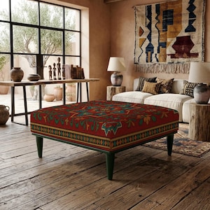 Op de afbeelding: Een vierkante poef met een felrode stoffen bekleding voorzien van een veelkleurig geometrisch en bloemmotief. Het meubelstuk rust op vier donkergroene taps toelopende poten en heeft een decoratieve rand langs de onderkant.