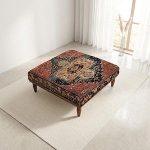 Puede incluir: Un otomano cuadrado con un intrincado patrón floral en azul oscuro, crema y naranja quemado en la parte superior y los laterales. La pieza se apoya sobre cuatro patas de madera cortas y cónicas que elevan la estructura tapizada.
