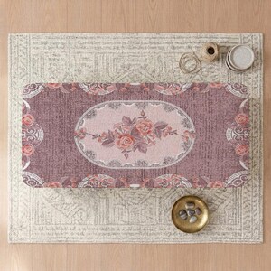 May include: A rectangular rug featuring a dusty rose and mauve color palette with a central oval medallion containing a detailed floral rose motif. The border displays intricate white lace-like patterns and smaller rose accents throughout the design.